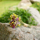 A small bouquet of yellow, purple, and white wildflowers tied with twine, resting on a large gray stone in an outdoor garden setting with blurred green grass and rocks in the background.