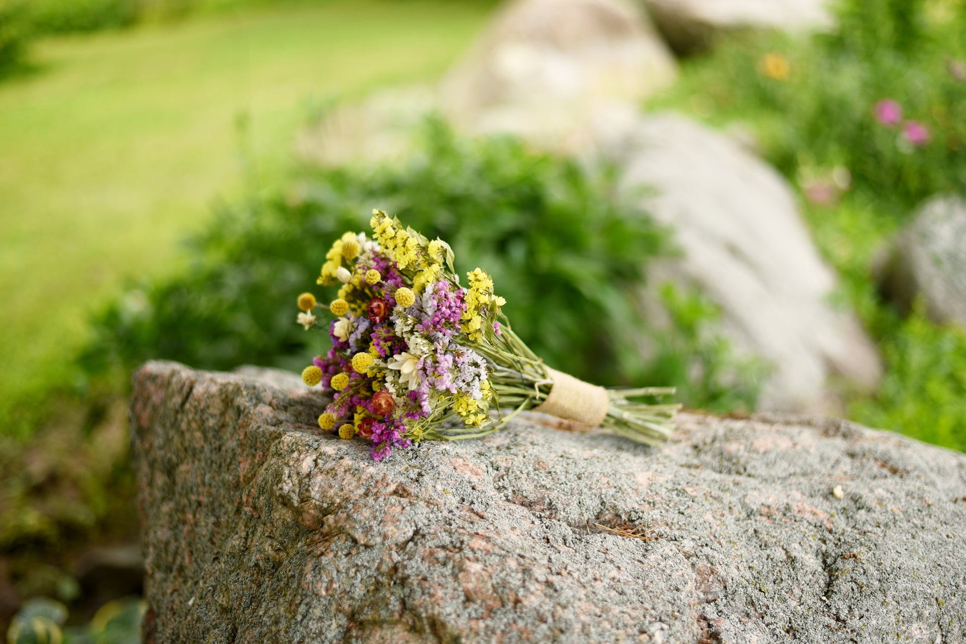 A small bouquet of yellow, purple, and white wildflowers tied with twine, resting on a large gray stone in an outdoor garden setting with blurred green grass and rocks in the background.