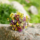 A small bouquet of dried multicolor flowers, including yellow, purple, white, and orange blossoms, resting on a textured gray stone with a blurred green garden background.