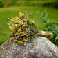 A small dried flower bouquet tied with a tan band rests on a large mossy rock in an outdoor garden setting with green grass and plants in the background.