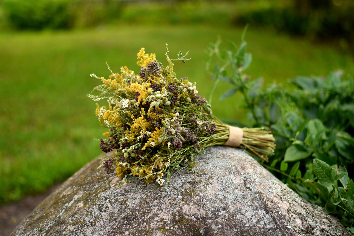 A small dried flower bouquet tied with a tan band rests on a large mossy rock in an outdoor garden setting with green grass and plants in the background.