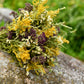 A small dried flower bouquet tied with a tan paper band, resting on a large gray stone with a blurred green outdoor background.