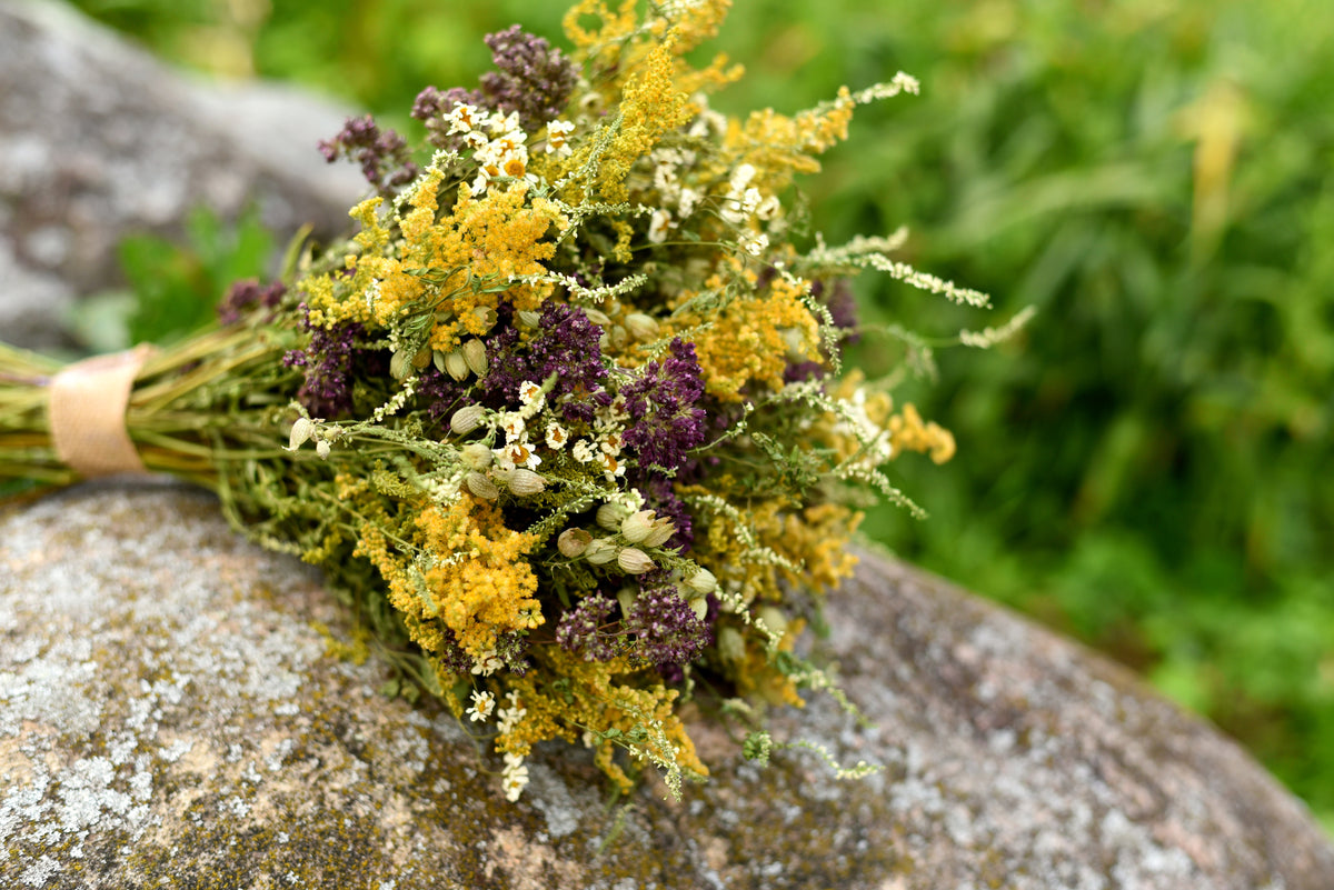 A small dried flower bouquet tied with a tan paper band, resting on a large gray stone with a blurred green outdoor background.