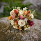 A small bouquet of dried flowers in cream, peach, pink, orange, and burgundy rests on a large mossy rock with a blurred natural background.