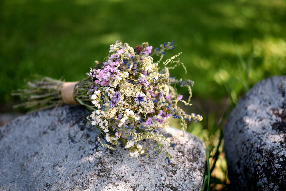 A small dried flower bouquet with white, purple, and blue blossoms tied with a tan band, resting on a gray stone outdoors with blurred green grass in the background.