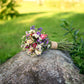 A small wildflower bouquet with purple, pink, white, and yellow blossoms tied with a tan ribbon, resting on a large mossy rock in an outdoor garden setting with green grass and foliage in the background.