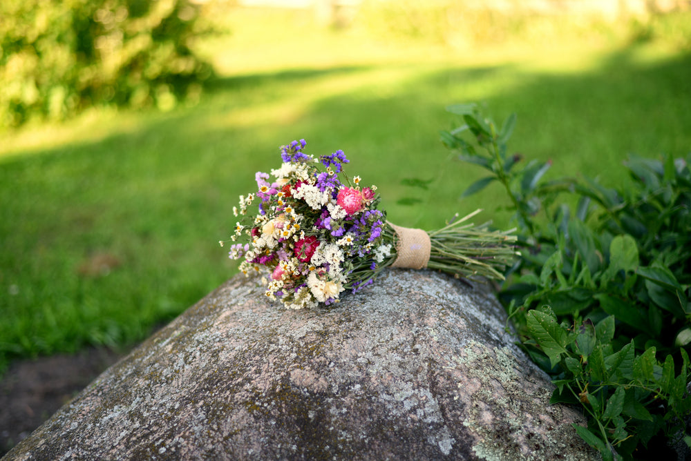 A small wildflower bouquet with purple, pink, white, and yellow blossoms tied with a tan ribbon, resting on a large mossy rock in an outdoor garden setting with green grass and foliage in the background.