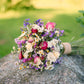 A small wildflower bouquet with pink, purple, white, and yellow flowers tied with a tan ribbon, resting on a large gray stone outdoors with a blurred green lawn and plants in the background.