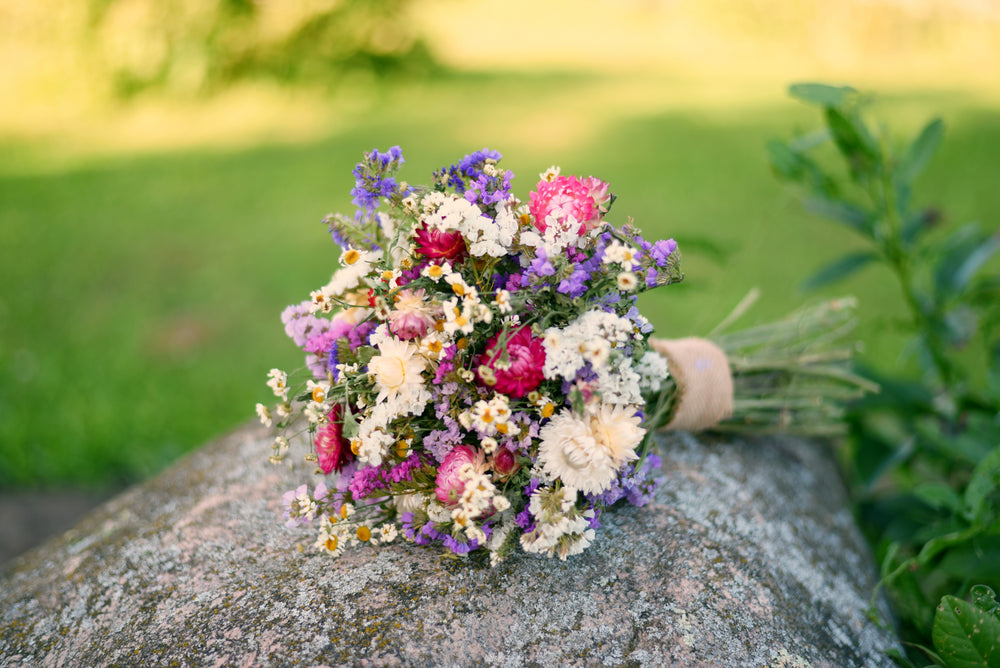 A small wildflower bouquet with pink, purple, white, and yellow flowers tied with a tan ribbon, resting on a large gray stone outdoors with a blurred green lawn and plants in the background.